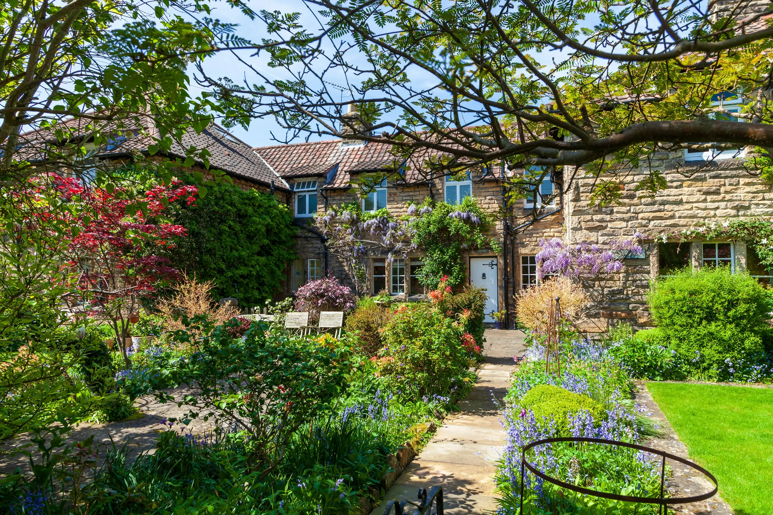 A lush spring garden with flowering borders and a stone path leading to a wisteria-covered house